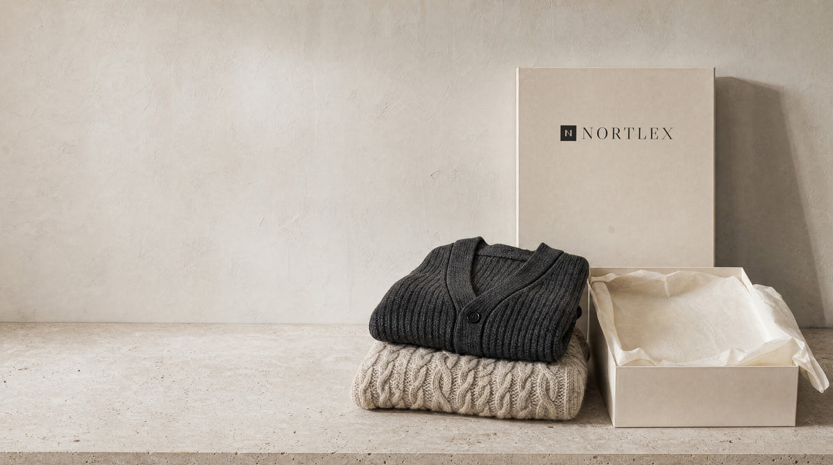 A folded charcoal ribbed cardigan and oatmeal cable-knit jumper on a limestone table beside an open Nortlex box lined with tissue paper